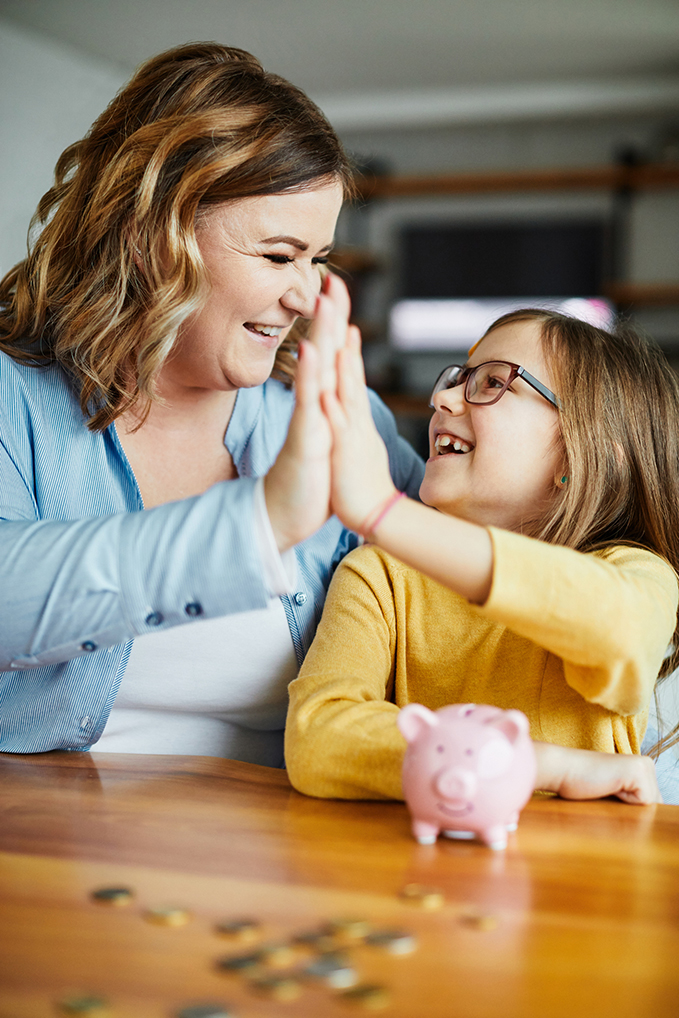 child smiling and giving adult a high-five with a piggy bank and coins in the foreground.